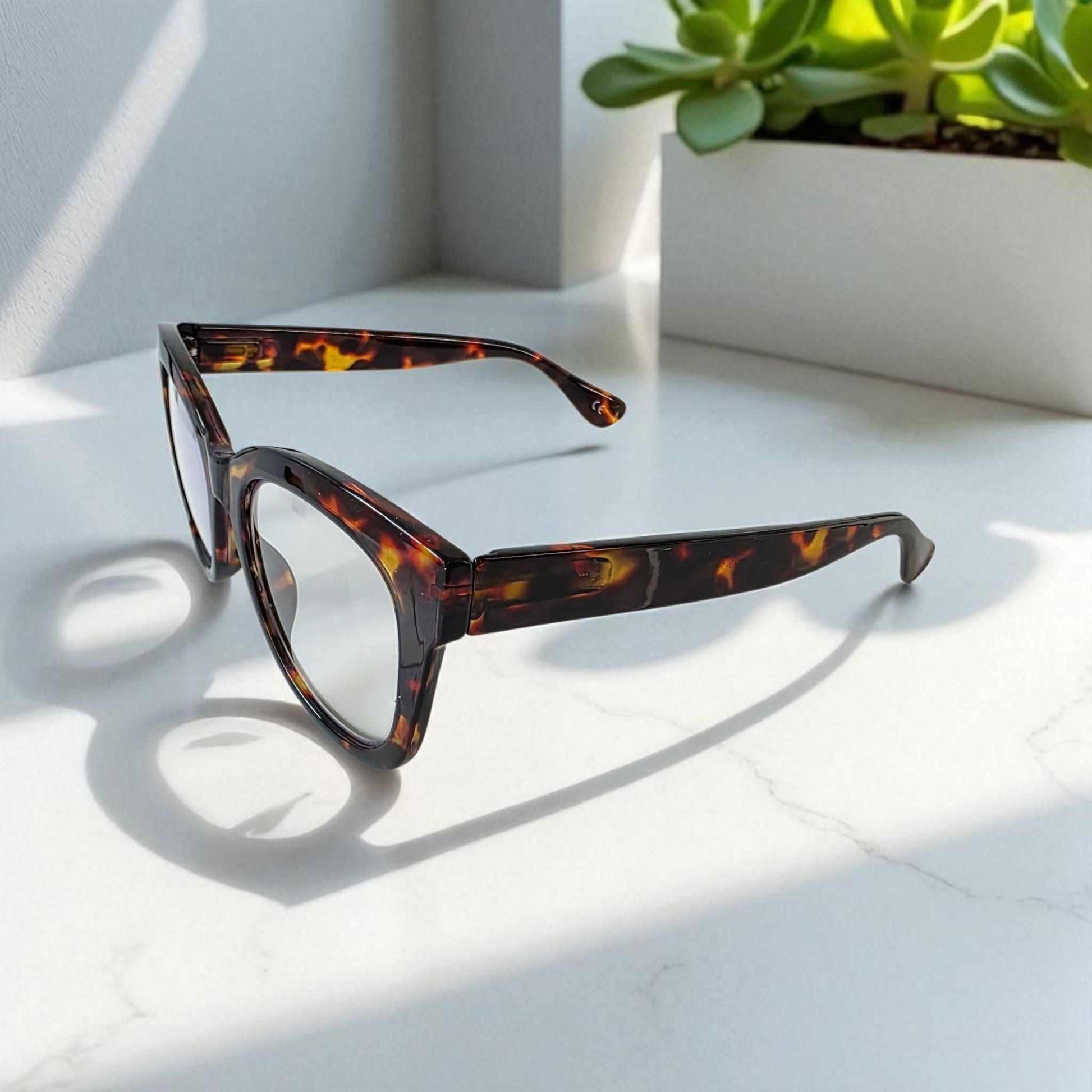 Bold tortoiseshell cat eye reading glasses with blue light filter on white surface near a green plant